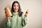 Young brunette woman holding dry italian macaroni over brown isolated background screaming proud and celebrating victory and