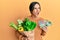 Young brunette woman holding groceries bag and australian dollars banknotes angry and mad screaming frustrated and furious,