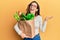 Young brunette woman holding paper bag with bread and groceries celebrating victory with happy smile and winner expression with