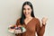 Young brunette woman holding plate with cake slices pointing thumb up to the side smiling happy with open mouth