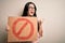 Young brunette woman holding protest banner with prohibited symbol over isolated background pointing and showing with thumb up to