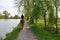 A young brunette woman in jeans and blue hoodie rides a bay horse along a path by the lake in the shade of a trees. Back view