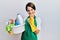 Young brunette woman with short hair wearing apron holding cleaning products beckoning come here gesture with hand inviting