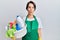 Young brunette woman with short hair wearing apron holding cleaning products making fish face with lips, crazy and comical gesture