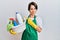 Young brunette woman with short hair wearing apron holding cleaning products pointing aside worried and nervous with forefinger,