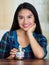 Young brunette woman sitting down facing camera, holding glass jar with coins inside, label reading vacations, smiling