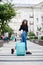 Young brunette woman, wearing black shirt, jeans and white hat with green sunglasses, holding mint luggage, walking in front of