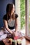 Young brunette woman, wearing black top and light pale pink yoga pants, sitting on windowsill with colorful aroma candles, looking