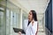 Young brunette woman, wearing white suit, holding black folder with documents,standing in white office building with huge windows.