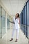 Young brunette woman, wearing white suit, holding black folder with documents,standing in white office building with huge windows.