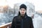 Young brutal man with a beard in a black stylish hat in a winter jacket on the background of winter trees in the city. Attractive