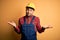 Young builder man wearing construction uniform and safety helmet over yellow isolated background clueless and confused expression