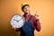 Young builder man wearing safety helmet holding big clock over yellow background surprised with an idea or question pointing