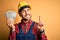 Young builder man wearing safety helmet holding dollars as payment over yellow background surprised with an idea or question