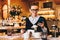 Young business woman in glasses sits in cafe at table, uses smartphone. On table is cup of coffee. Girl working