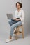 Young business woman posing isolated over grey wall background sitting on stool using laptop computer