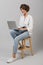 Young business woman posing isolated over grey wall background sitting on stool using laptop computer