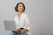 Young business woman posing isolated over grey wall background sitting on stool using laptop computer