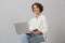 Young business woman posing isolated over grey wall background sitting on stool using laptop computer