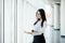 Young business woman working in her luxurious office holding a laptop standing against panoramic window with a view on business di