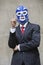 Young businessman in pinstripes suit and wrestling mask pointing up over gray background