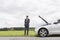 Young businessman standing arms crossed by broken down car at countryside