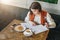 Young businesswoman is sitting in cafe at table, signing documents. Student is studying online, doing homework.