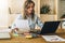 Young businesswoman woman is sitting at kitchen table and uses laptop, working, studying. On table tablet computer