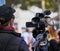 Young camerawoman in blue anorak with cap and sunglasses setting up her video camera mounted on a tripod