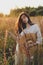 Young carefree female in rustic linen cloth posing in summer meadow. Summer delight and travel. Stylish boho woman with straw hat