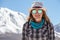 Young casual blond woman in hat and sunglasses looking at camera with snow covered mountains on the background