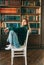 Young caucasian cheerful woman student sitting in a white chair near bookshelf in library