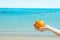 Young Caucasian Female Hand Holding Whole Ripe Orange on Blue Sky Turquoise Sea Background. White Sand Waves. Summer Vacation