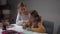 Young Caucasian female tutor teaching cute little girl at home. Smart schoolchild writing in the exercise book, teacher