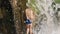 A young Caucasian girl in a blue swimsuit walks through a large wall of falling water on a mountain waterfall. A woman