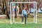 Young Caucasian goalkeeper girl with flushed face posing by outdoor football goal with foot put on ball looking into camera