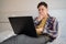 Young caucasian male remote worker with a laptop sits at home during the day on the bed in casual clothes, covers his