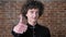 Young caucasian man with curly hair showing thumbs up into camera and smiling, confident male, brick wall background