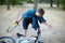 Young caucasian man with dark hair prepares to saw the bicycle laying on the ground in abandoned park with big blue and yellow han