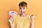 Young caucasian man holding 100 new taiwan dollars banknotes pointing thumb up to the side smiling happy with open mouth