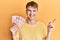 Young caucasian man holding 100 new taiwan dollars banknotes smiling happy pointing with hand and finger to the side