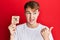 Young caucasian man holding under 18 prohibition sticker pointing thumb up to the side smiling happy with open mouth