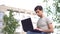 A young caucasian man with sits on the grass near the house and typing on a laptop. Remote work