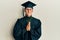 Young caucasian man wearing graduation cap and ceremony robe praying with hands together asking for forgiveness smiling confident
