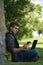 Young caucasian man working on his computer sitting under a tree while he is looking at his mobile at the same time. Technological