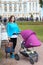 Young Caucasian mother with stroller walking in summer park near palace, excursion in Russian historical places