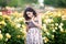 Young Caucasian woman with dark curly hair near yellow rose bush in a rose garden looking to her black smartphone, reading emails,