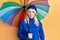 Young caucasian woman holding colorful umbrella in shock face, looking skeptical and sarcastic, surprised with open mouth