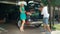 Young Caucasian woman leans on the car while husband stuffs the trunk with bags