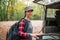 A young Caucasian woman poses with a backpack on her back and closes the trunk of a jeep. In the background, trees and forest.